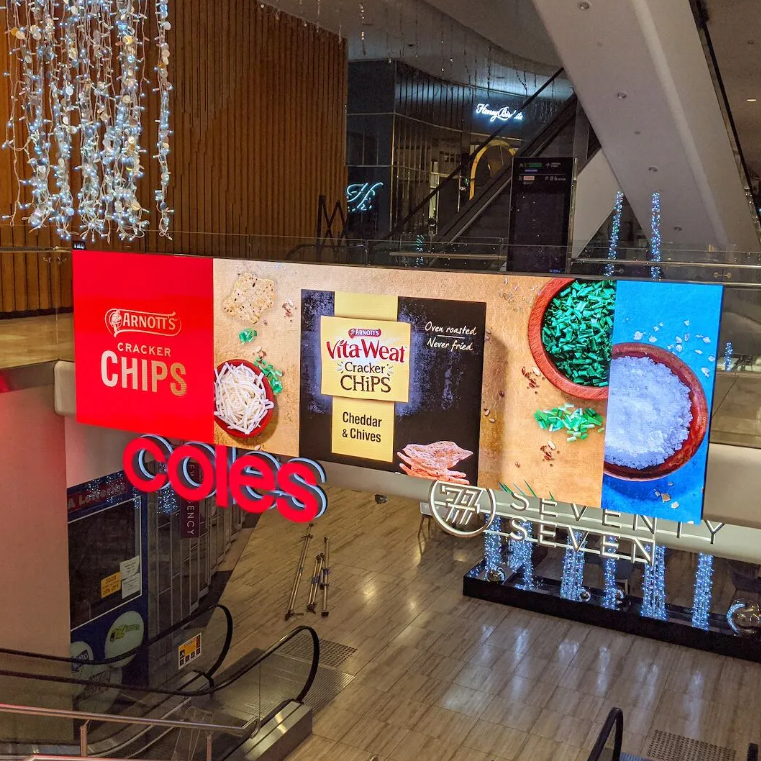 Indoor mall atrium with a bright digital ad panel featuring Arnott's chips and VitaWeat Cheddar & Chives, and a prominent Coles sign.