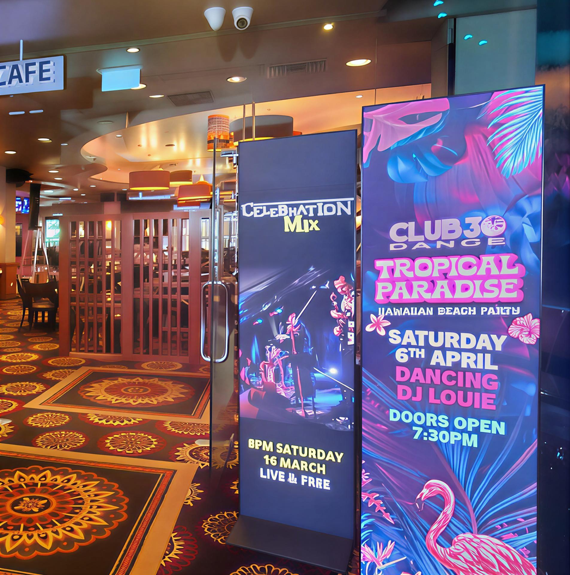 Entrance of a vibrant entertainment venue with large colorful banners promoting Tropical Paradise, Club 30, and Celebration Mix banners inside a casino-like lobby.