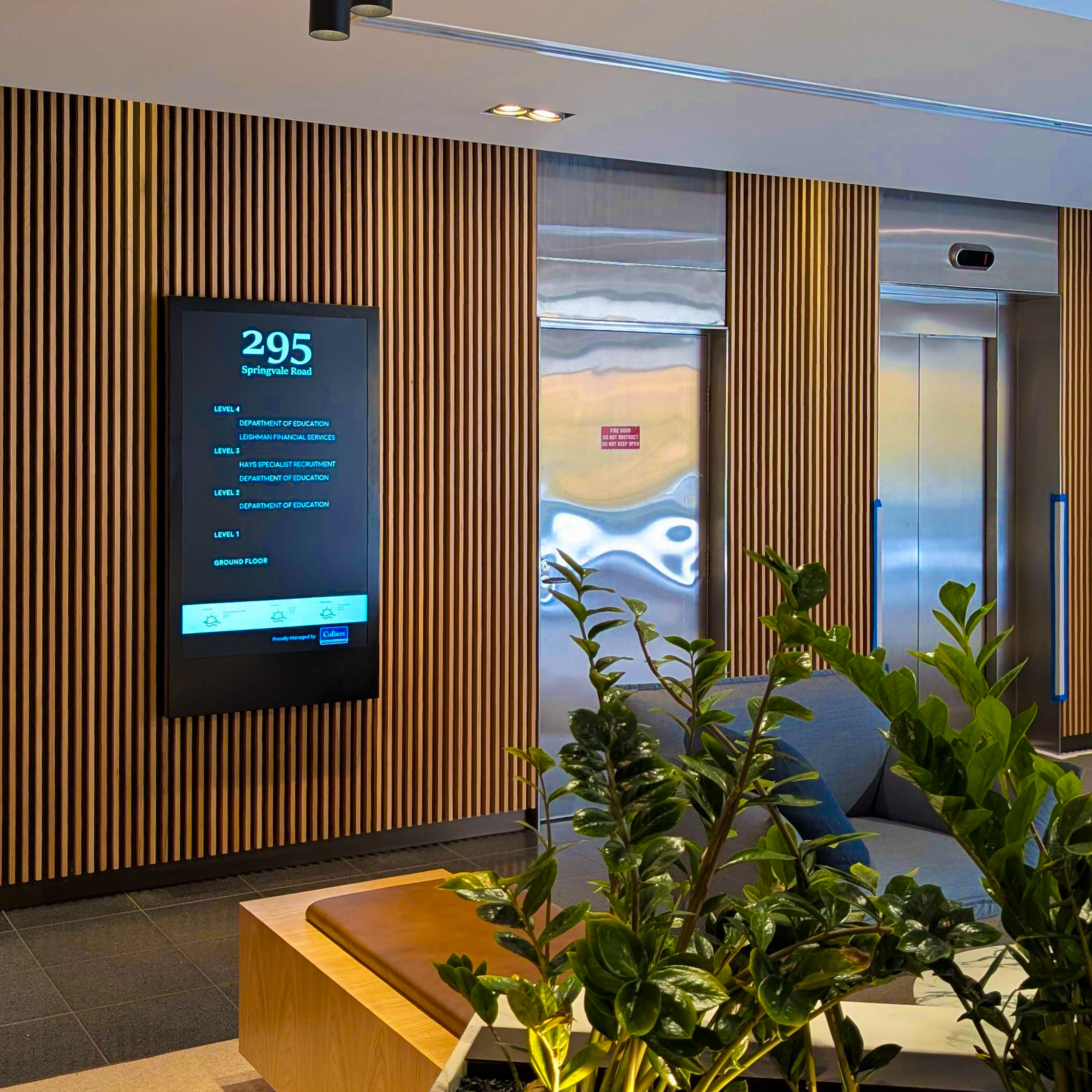 Interior lobby with vertical wooden slat walls, a digital directory screen showing 295 Springvale Road, and glass elevators in the background, plus green plants in the foreground.