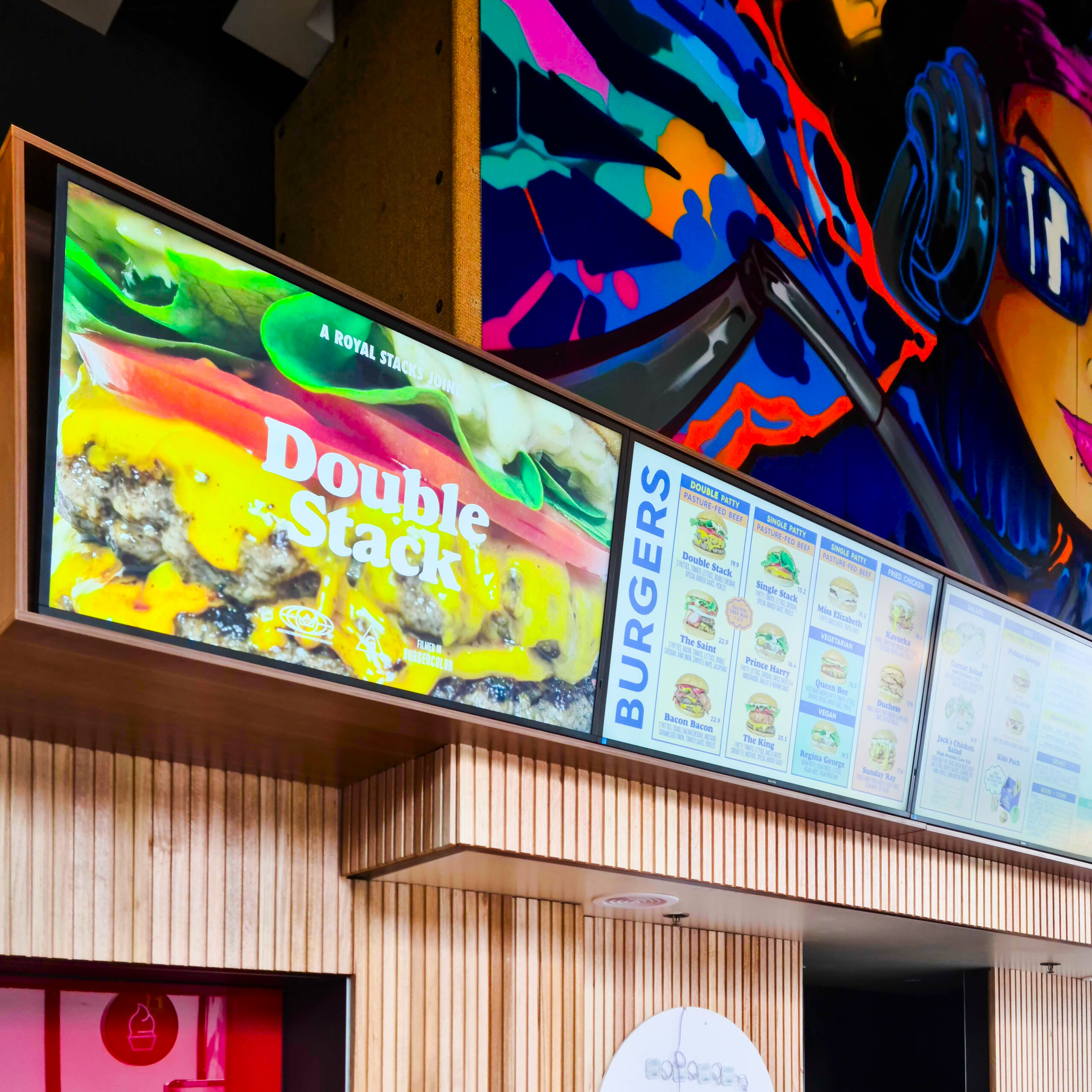 Colorful abstract mural above angled digital menu boards displaying burgers in a fast-food restaurant interior.