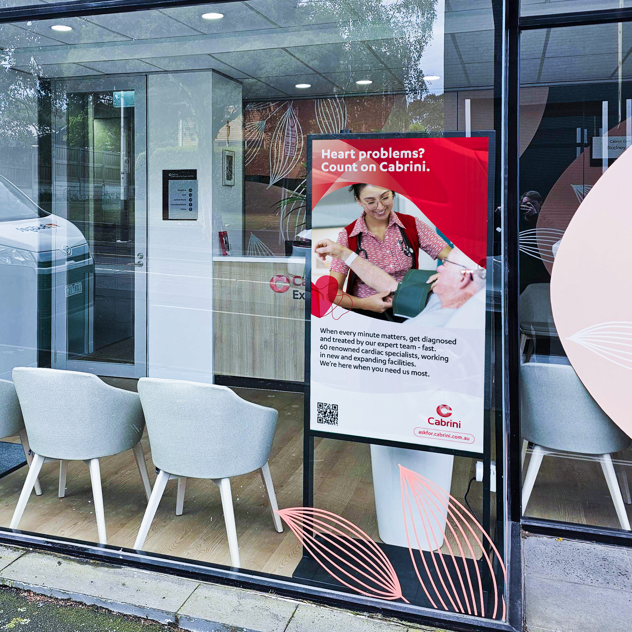 Storefront window with pink curved graphic posters, gray chairs outside, and reflections in glass; interior ads for Cabrini healthcare.