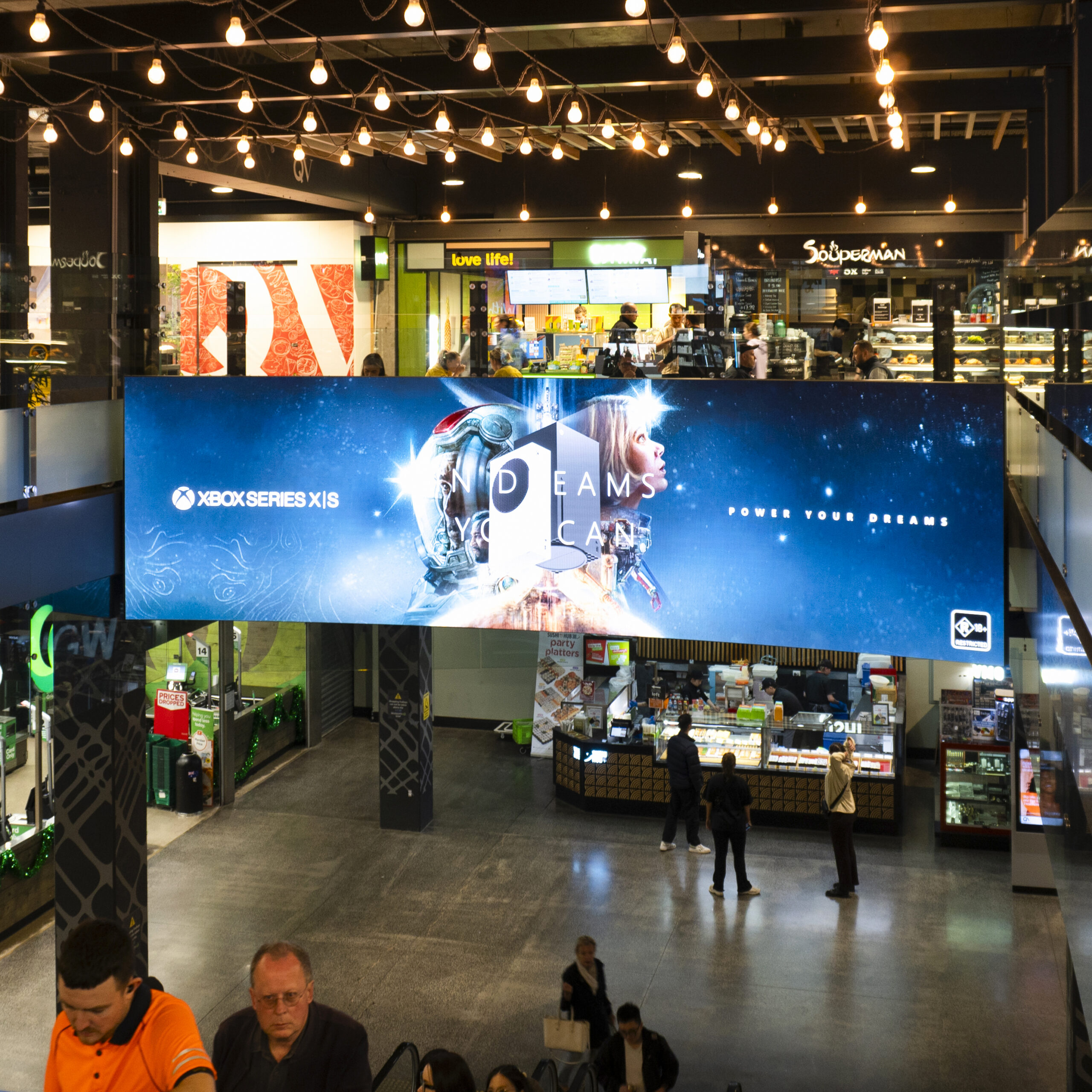 Indoor shopping center with a large Xbox banner, storefronts in the background, and people walking beneath string lights.