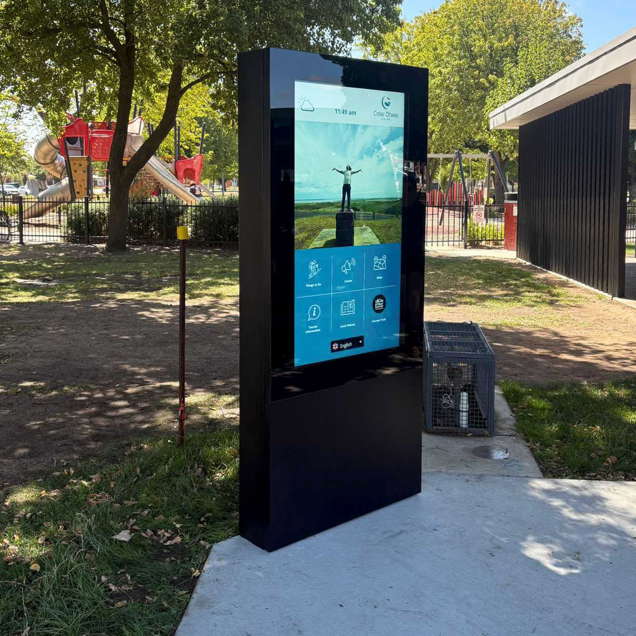 Outdoor public information kiosk with a large touchscreen in a park beside a playground; screen shows a person with arms raised and blue icon grid.