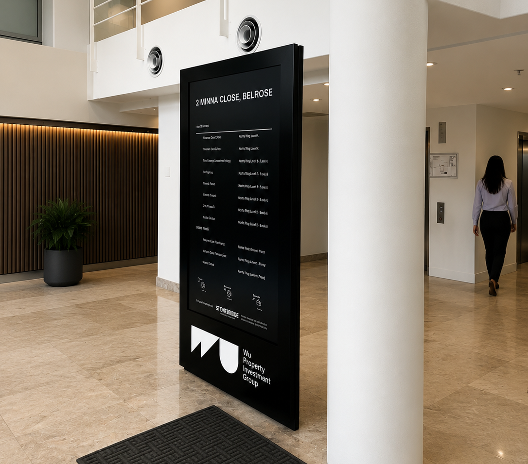Black directory board in a bright modern lobby with a white column and railing above; a person walks toward an elevator on the right.