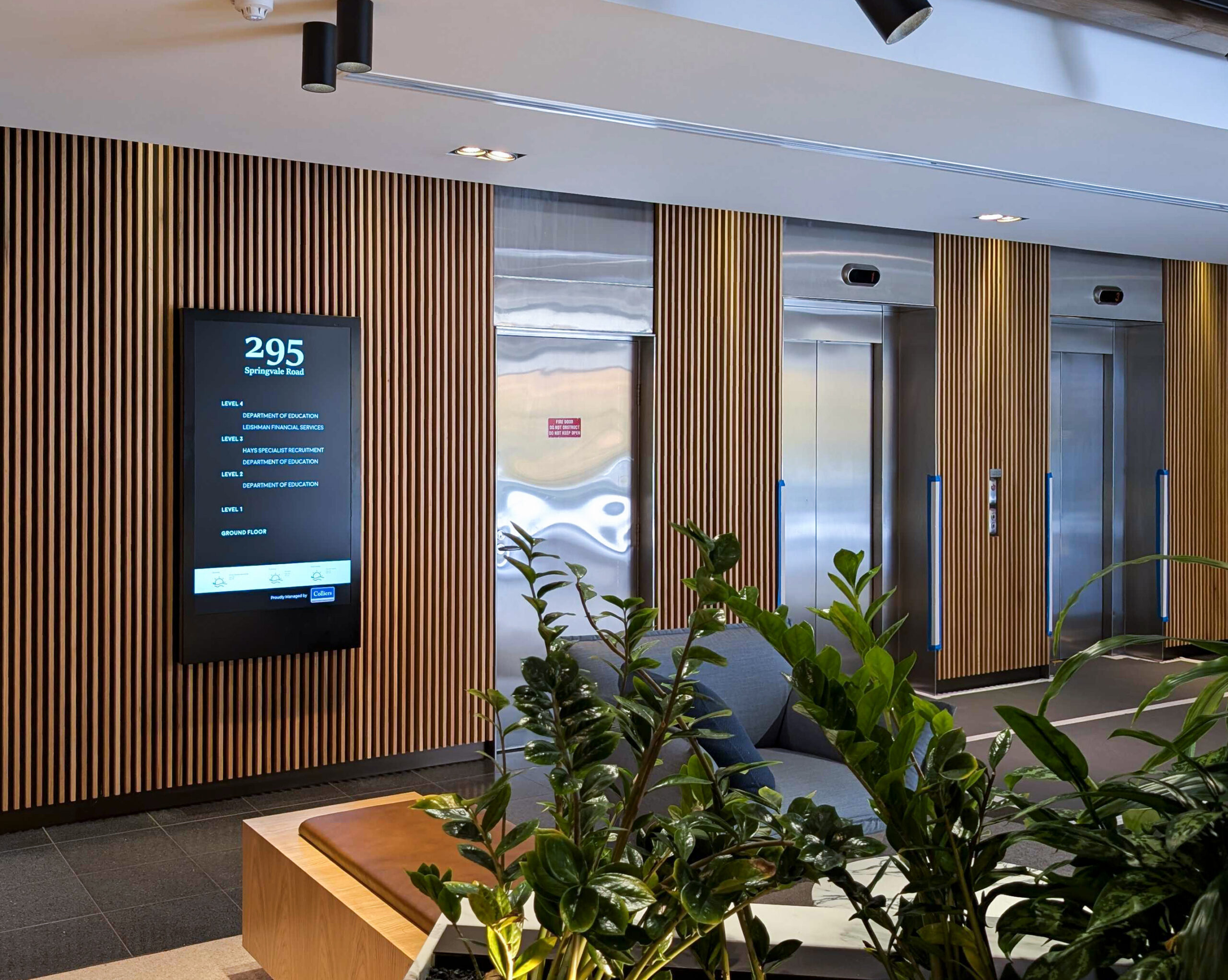 Interior lobby with vertical wooden slat walls, metal elevator doors, a digital directory screen, and potted plants.