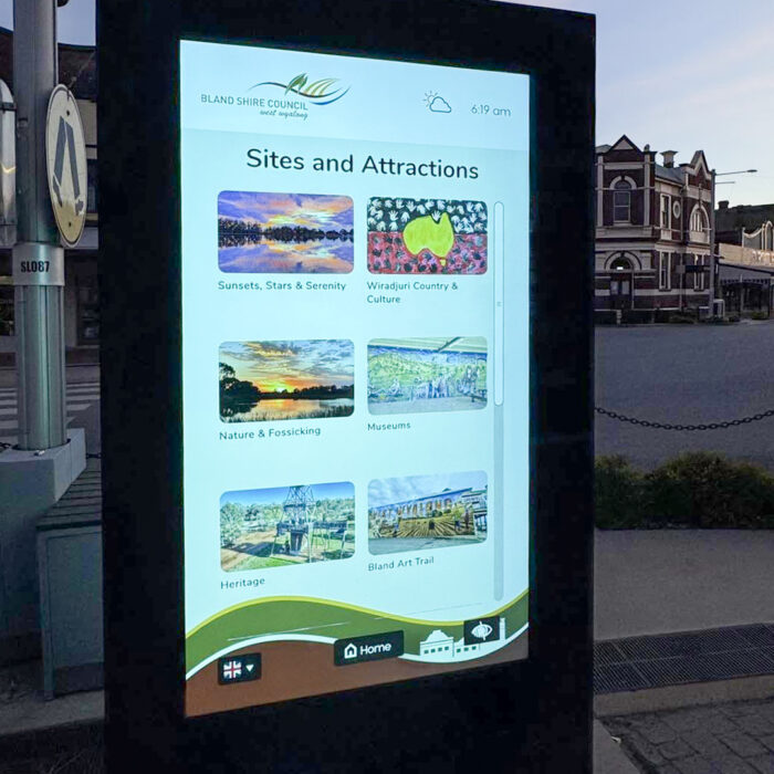 Outdoor digital information kiosk displaying a grid of local sites and attractions (Sites and Attractions) with categories like Sunsets, Nature, Museums; sign shows time and weather icons.