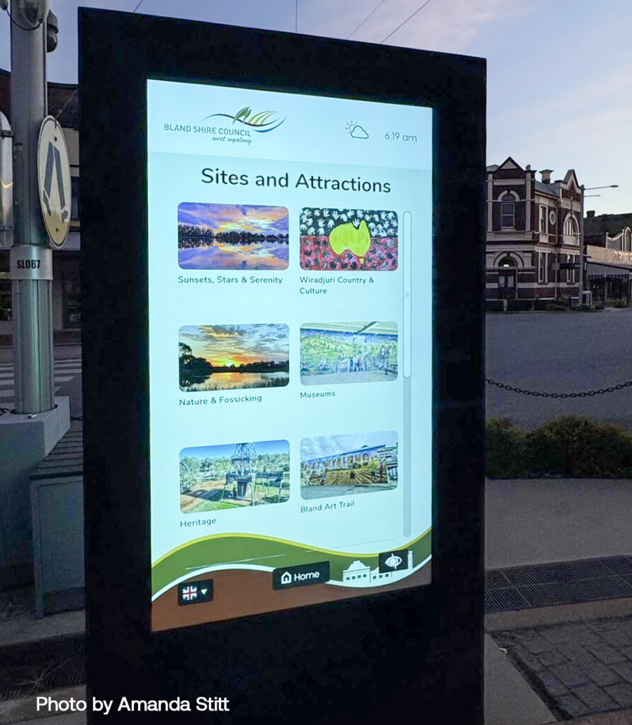 Outdoor digital information kiosk displaying a grid of local sites and attractions (Sites and Attractions) with categories like Sunsets, Nature, Museums; sign shows time and weather icons.