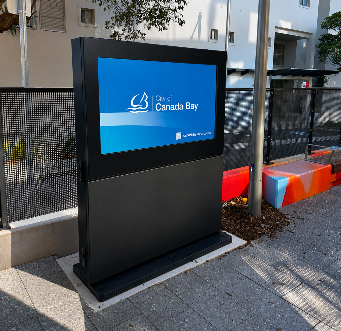Outdoor digital information kiosk displaying the City of Canada Bay logo on a blue screen, mounted on a black pedestal beside a mesh fence and colorful barrier.
