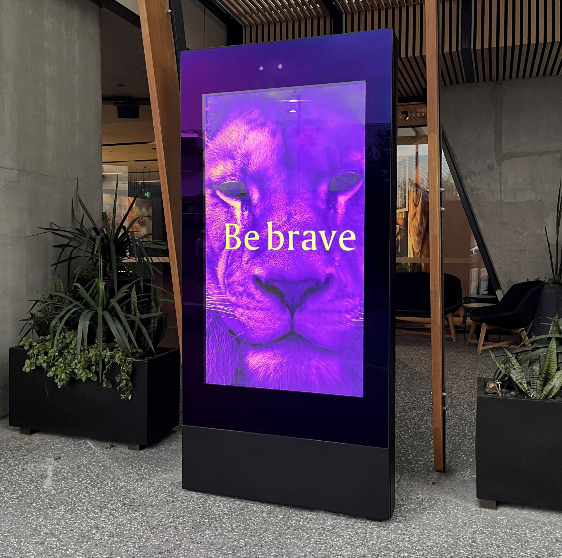 Digital advertisement panel with a purple lion close-up and the text 'Be brave' in yellow, set in a modern lobby with plants and wooden beams.