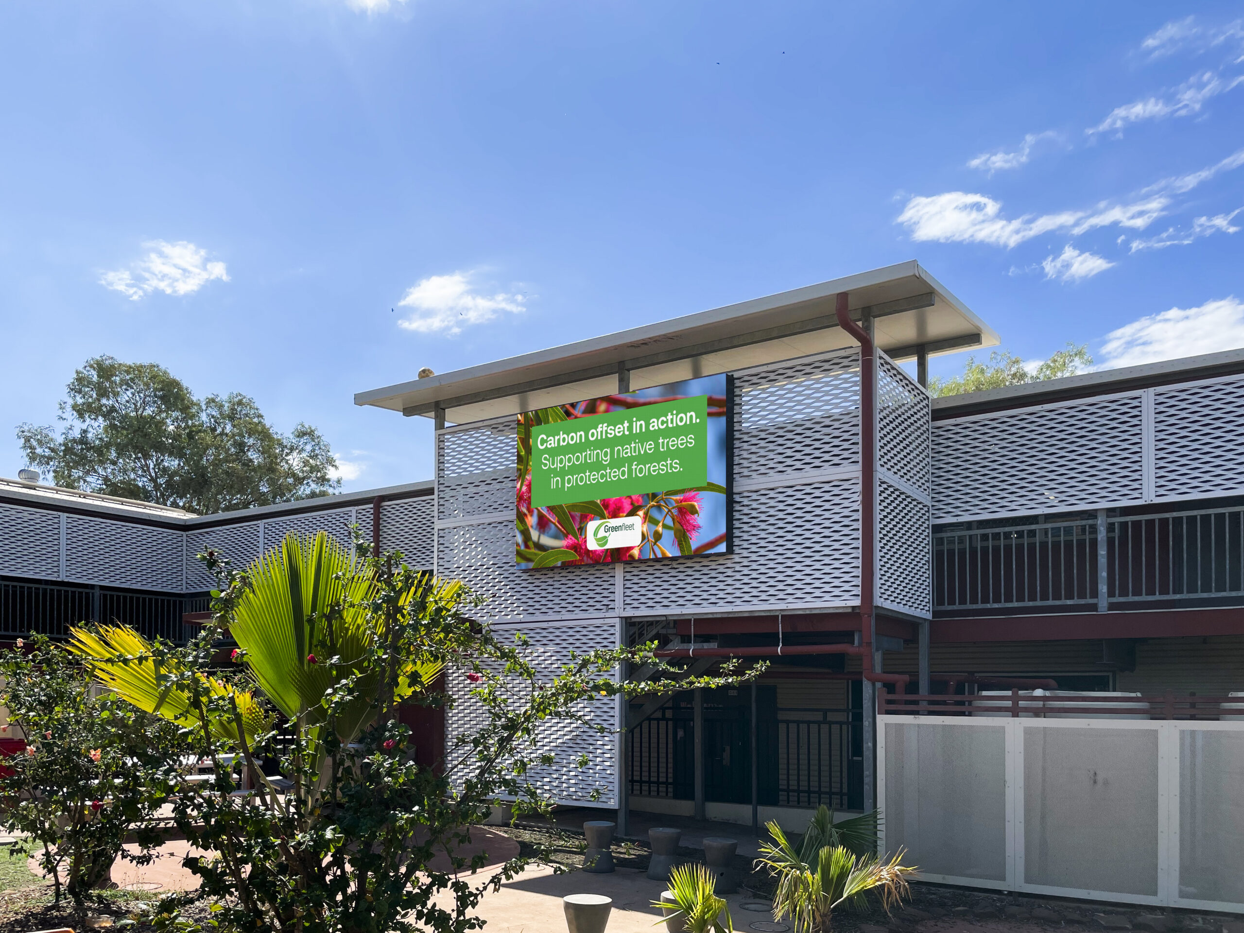 Green energy banner on a modern building reads: 'Carbon offset in action. Supporting native trees in protected forests.' with company logo, set against a blue sky and lattice walls.
