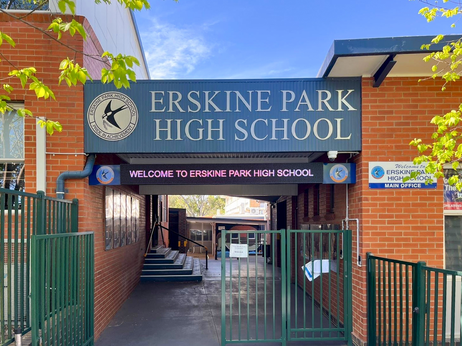 Entrance to Erskine Park High School with a large blue sign and brick walls, green gate, and trees overhead.