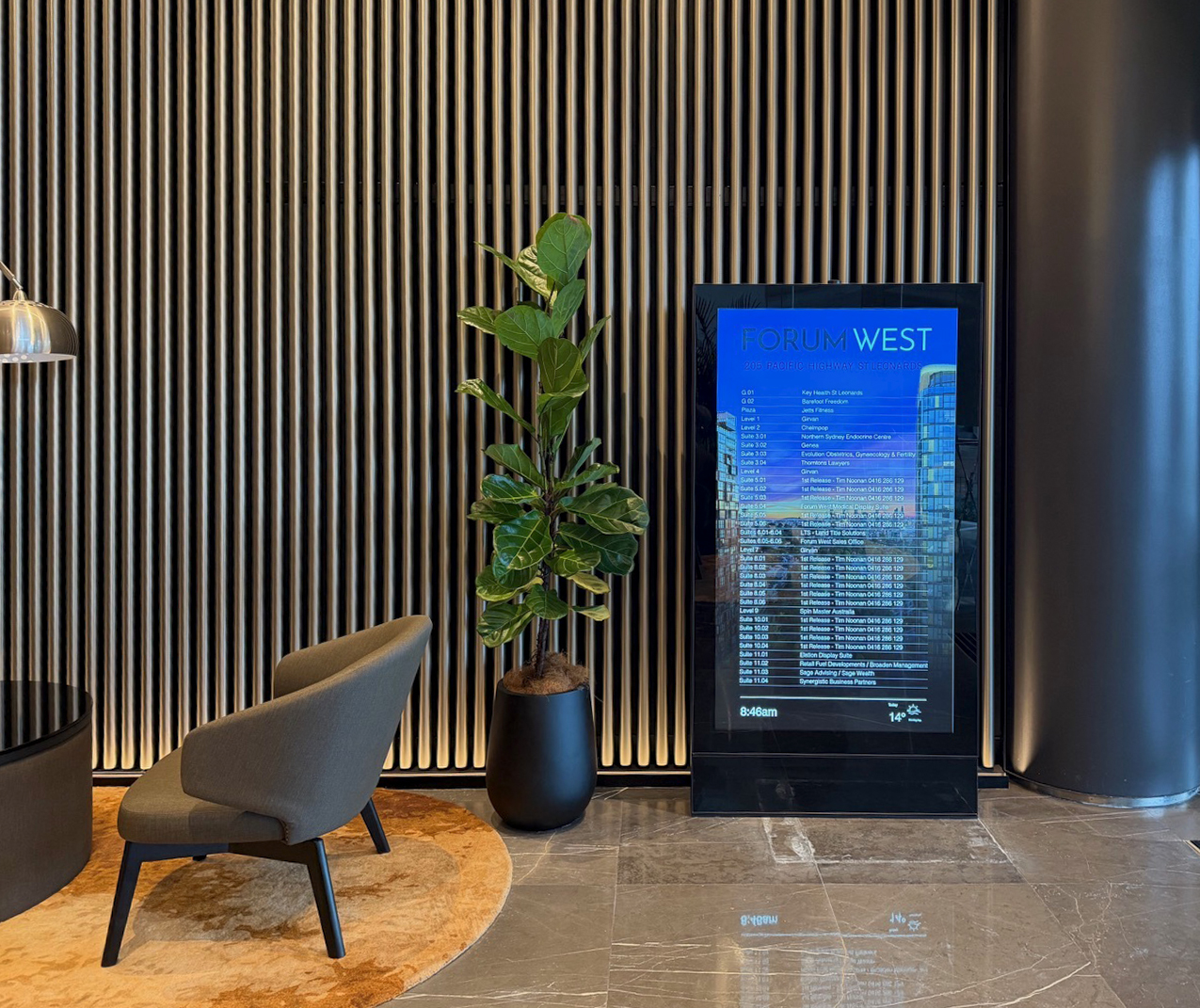 Modern lobby area with a tall green plant in a black pot, a gray lounge chair on a round orange rug, and a digital directory sign reading FORUM WEST.