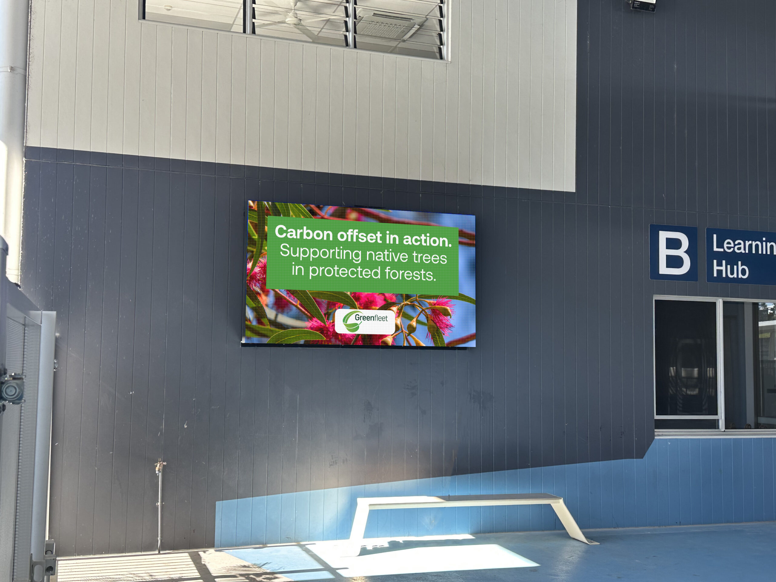Green fleet sign on dark wall: 'Carbon offset in action. Supporting native trees in protected forests.' with colorful background vegetation and Greenfleet logo above a doorway sign reading 'Learnin... Hub' in the corner.