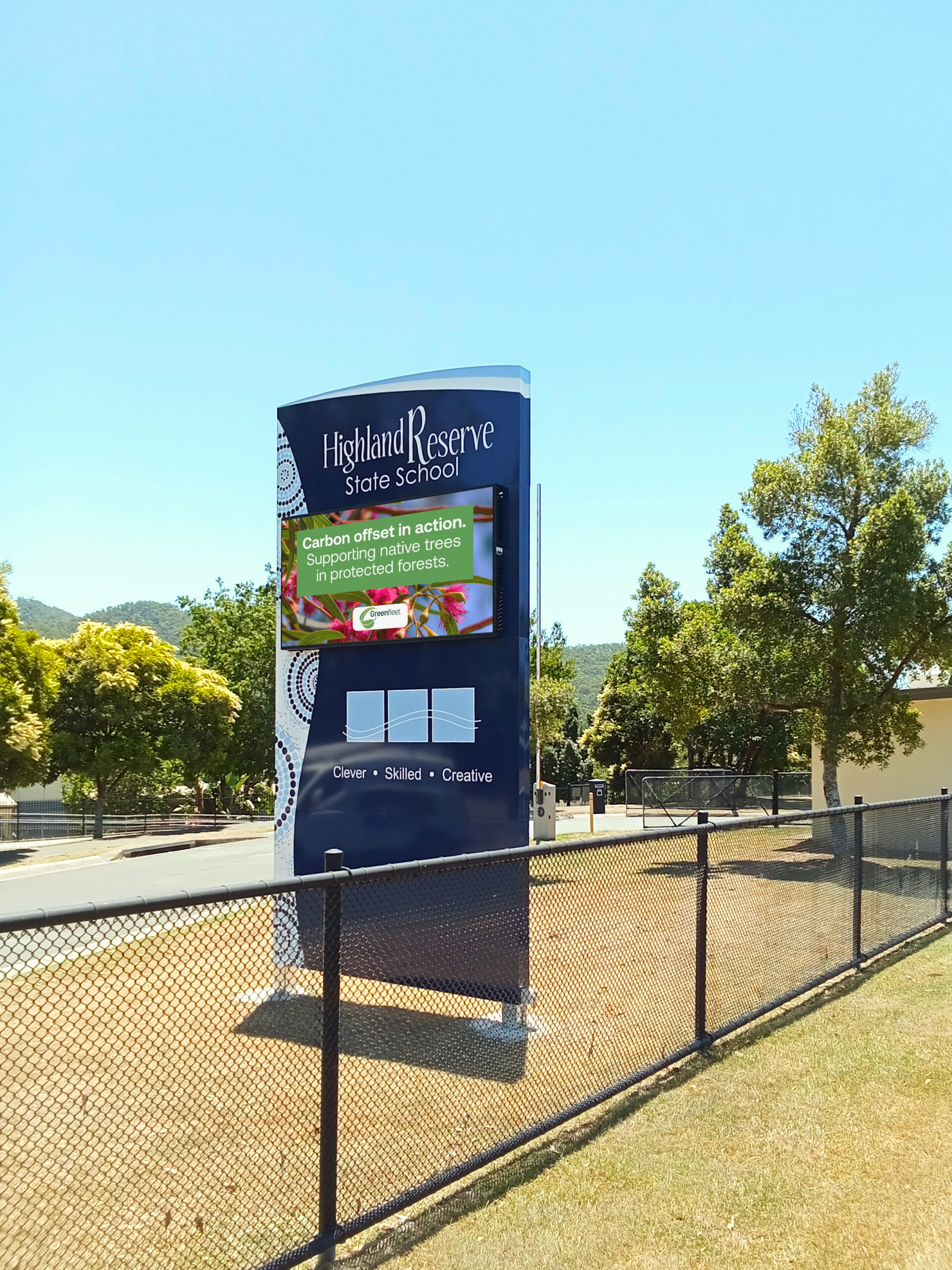 Blue Highland Reserve State School sign with a green carbon-offset message on a sunny day, next to a chain-link fence and trees.