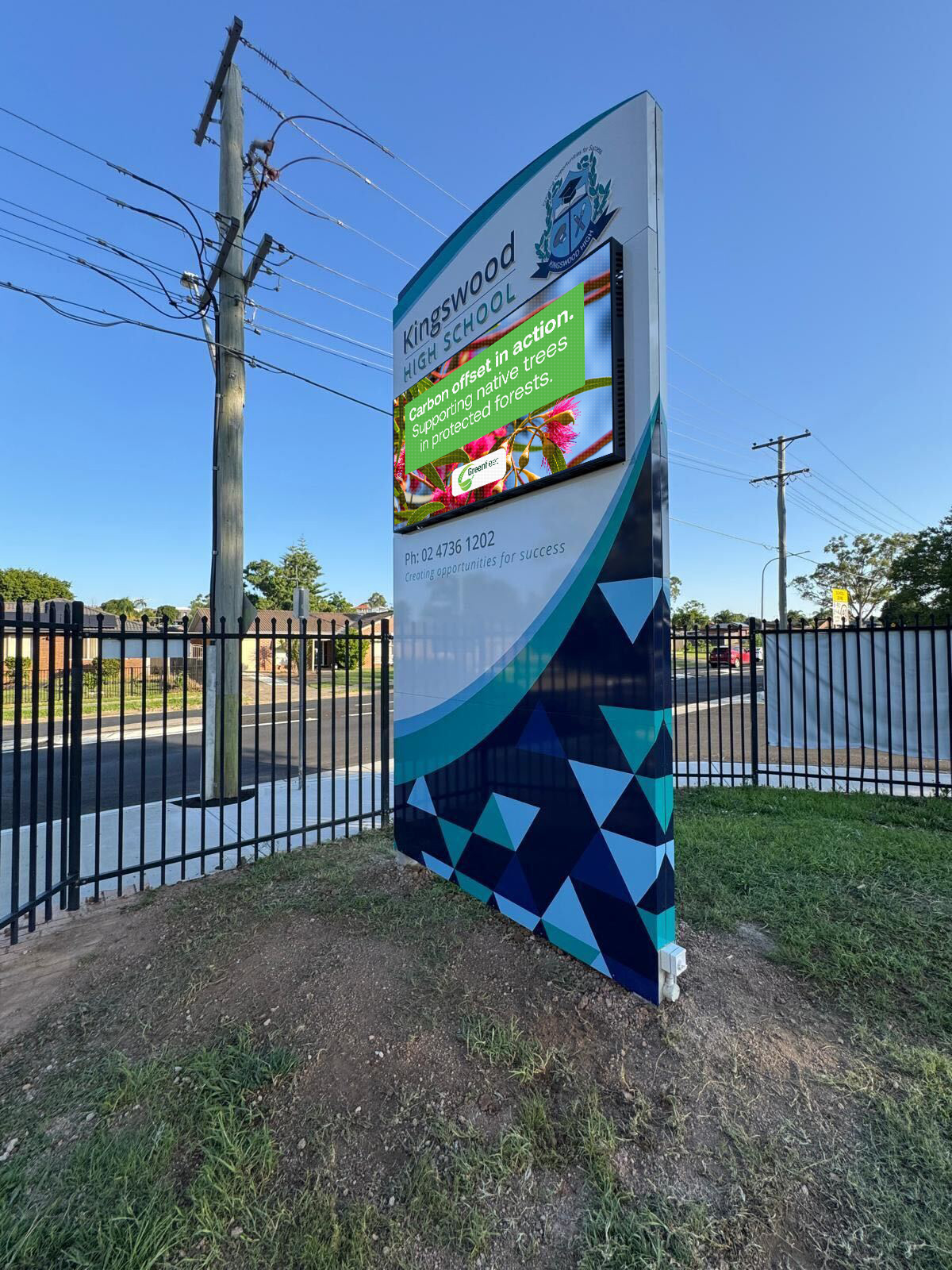 Tall school sign for Kingswood High School with a digital display, beside a fence and utility pole on a sunny street.