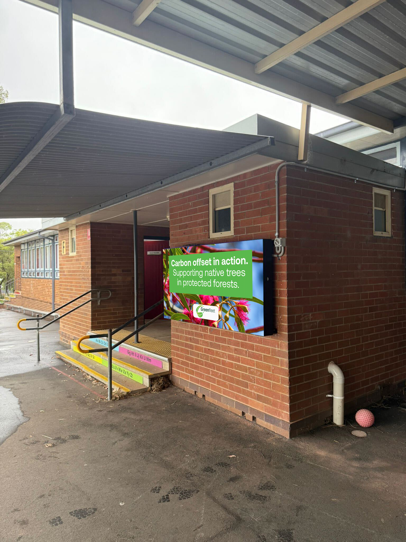 Brick building under a metal canopy with a bright sign about carbon offsetting and native trees, and colorful ramp/steps nearby.