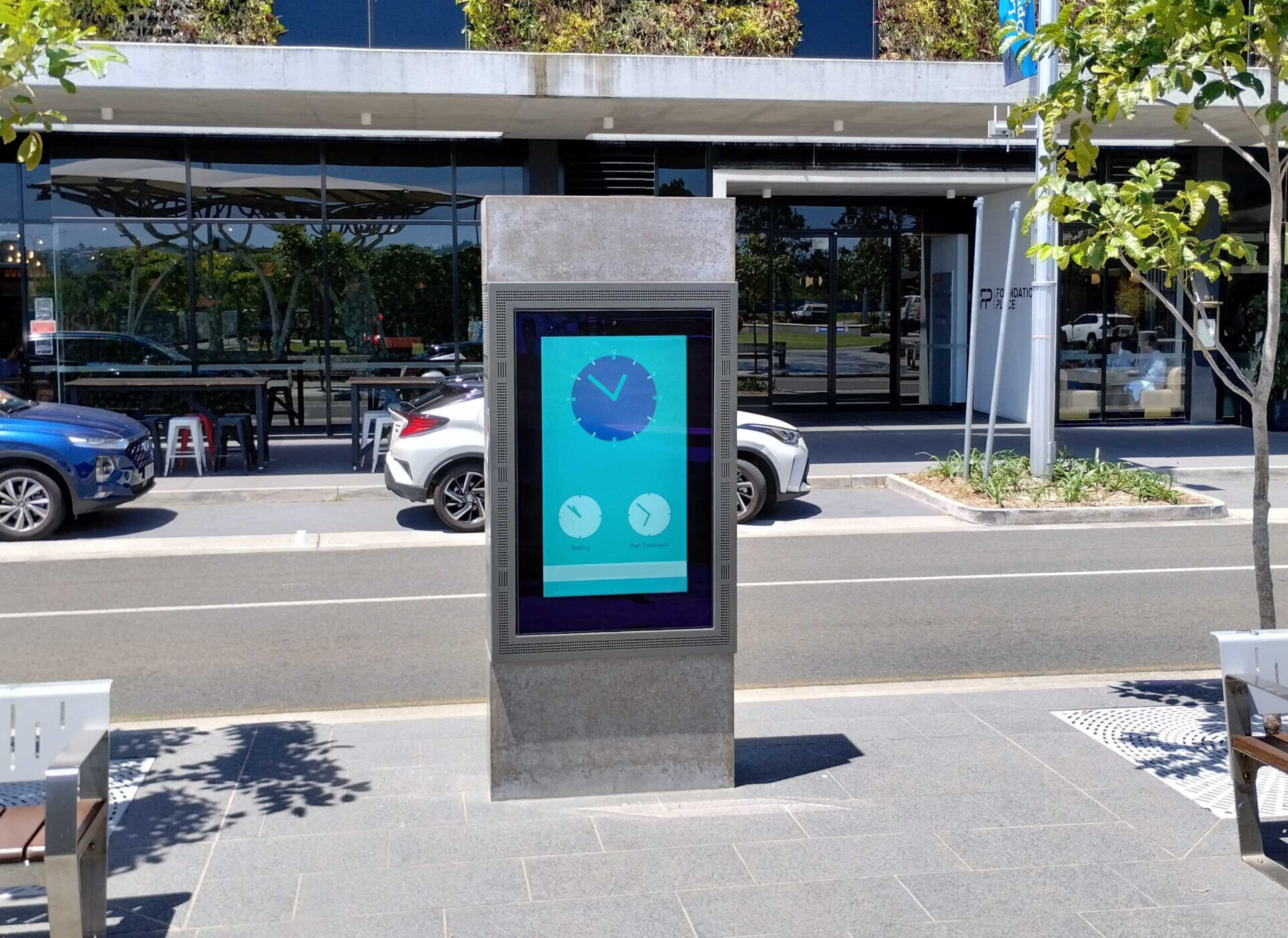 Public square with a tall digital information kiosk in the foreground, beside a modern building with green vertical gardens and blue panels.