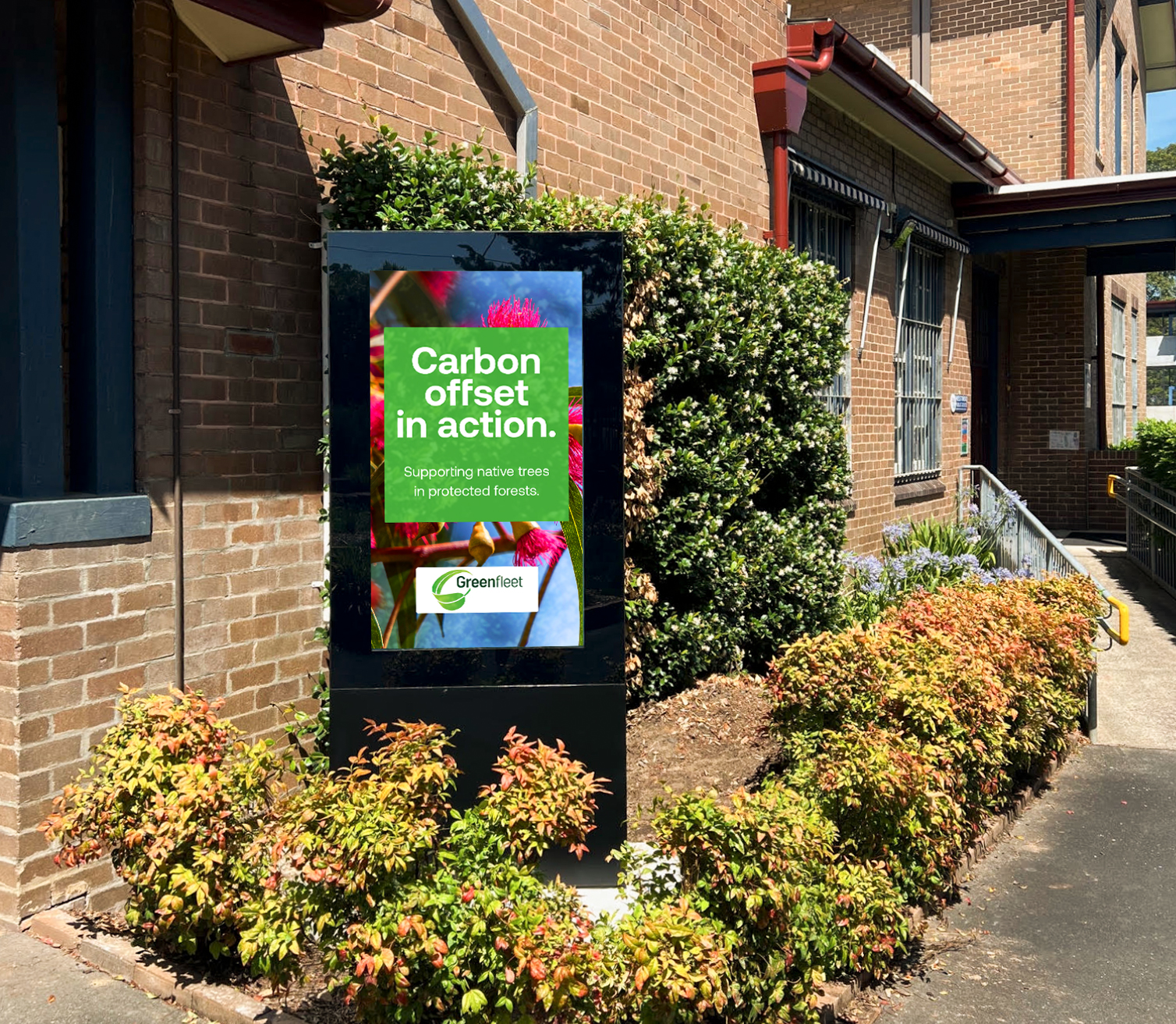 Poster on a freestanding display reading 'Carbon offset in action' by Greenfleet, outdoors beside shrubs and a brick building.