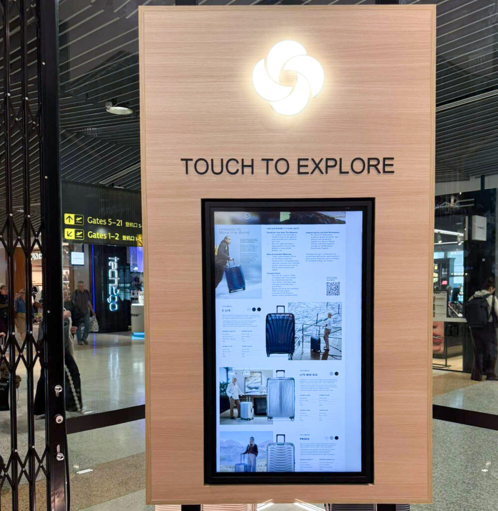 Wooden information kiosk with a touchscreen displaying luggage brochures; sign above reads 'TOUCH TO EXPLORE'.