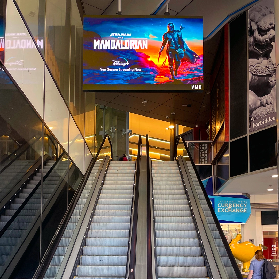 Indoor mall scene with mirrored escalators converging toward a large digital ad board showing The Mandalorian poster (Disney+).