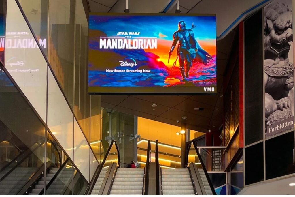 Large indoor escalators beneath a hanging digital screen advertising The Mandalorian on Disney+ in a mall.